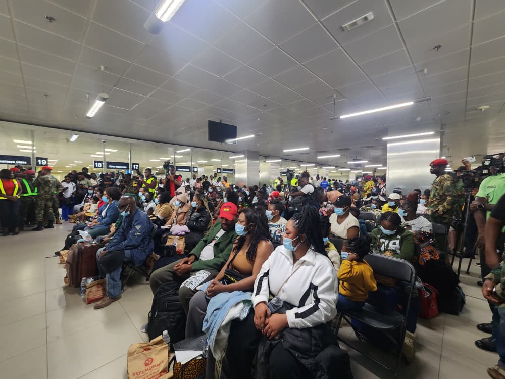 Crowd of travelers wearing masks seated in an airport waiting area with luggage, security personnel visible near counters in the background.