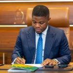 Professional man in a navy pinstripe suit signs documents at an executive desk in a modern office.