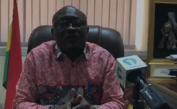 Screenshot-2026-04-30-155921-750x375 | Ghanaguardian.com Man in a red patterned shirt sits at a desk in an office and speaks into a microphone during a press briefing, with a flag and framed photo behind him.