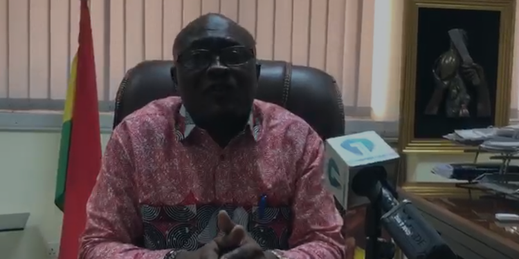 Man in a red patterned shirt sits at a desk in an office and speaks into a microphone during a press briefing, with a flag and framed photo behind him.