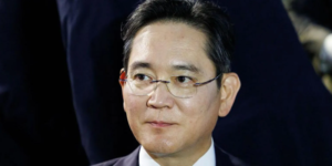 Man wearing glasses and a dark suit, looking to the side in a formal setting.