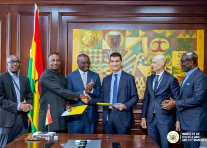 Six men in suits participate in a formal signing ceremony: two center figures shake hands and exchange documents, with a flag behind them and an official crest in the corner.