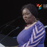 Woman in a blue dress with white chevron sleeves speaks at a podium; 3i Africa Summit logo visible in top right corner.