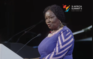 Woman in a blue dress with white chevron sleeves speaks at a podium; 3i Africa Summit logo visible in top right corner.