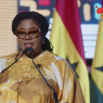 Female speaker at a podium wearing a gold and white patterned dress and brown glasses, with flags and a '3i Africa Summit' logo in the background.
