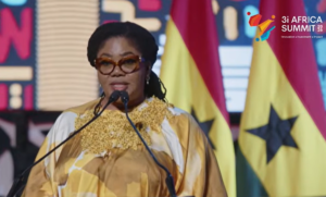 Female speaker at a podium wearing a gold and white patterned dress and brown glasses, with flags and a '3i Africa Summit' logo in the background.