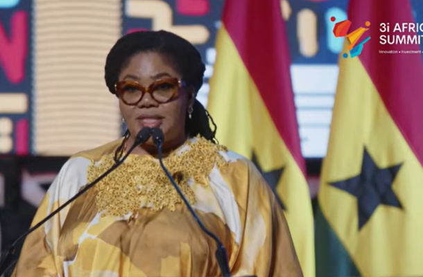 Female speaker at a podium wearing a gold and white patterned dress and brown glasses, with flags and a '3i Africa Summit' logo in the background.