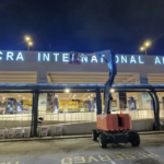 Nighttime outside Accra International Airport; a red cherry picker extends a platform near the terminal entrance under bright signlights.