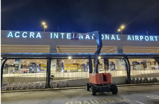 Nighttime outside Accra International Airport; a red cherry picker extends a platform near the terminal entrance under bright signlights.