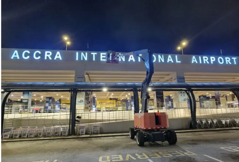 Nighttime outside Accra International Airport; a red cherry picker extends a platform near the terminal entrance under bright signlights.
