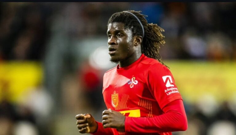 Soccer player in a red jersey sprinting on the field during a match near blurred stands.