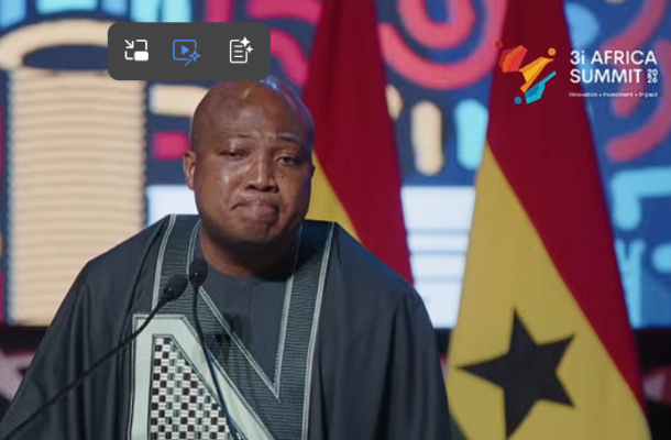 An African man in a dark agbada-style outfit speaks at a podium during a speech at the 3i Africa Summit, with flags and a colorful backdrop behind him.