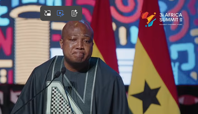 An African man in a dark agbada-style outfit speaks at a podium during a speech at the 3i Africa Summit, with flags and a colorful backdrop behind him.