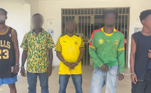 Five young men with blurred faces stand in a row outside a building, each wearing different casual or sports outfits including jerseys and patterned shirts.