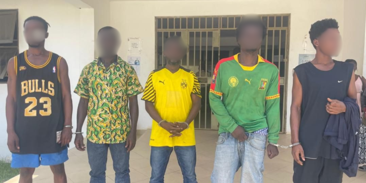 Five young men with blurred faces stand in a row outside a building, each wearing different casual or sports outfits including jerseys and patterned shirts.