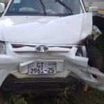 White compact SUV with front-end collision damage, crumpled hood and bumper, license plate GT 2961-25 visible, parked beside a larger truck in a dirt lot.