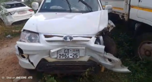 White compact SUV with front-end collision damage, crumpled hood and bumper, license plate GT 2961-25 visible, parked beside a larger truck in a dirt lot.