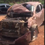 Brown SUV severely damaged at the front, with the hood crumpled and engine exposed, parked on a dirt lot.