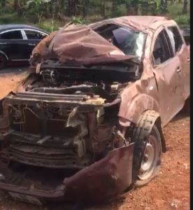 Brown SUV severely damaged at the front, with the hood crumpled and engine exposed, parked on a dirt lot.