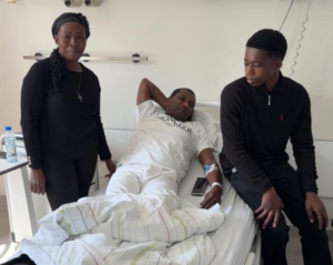 Man in a hospital bed with an IV, flanked by a standing woman and a young man sitting at the foot of the bed in a hospital room.