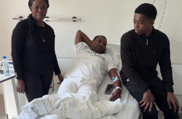 Man in a hospital bed with an IV, flanked by a standing woman and a young man sitting at the foot of the bed in a hospital room.