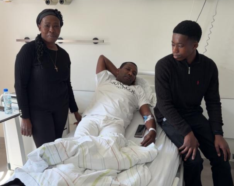 Man in a hospital bed with an IV, flanked by a standing woman and a young man sitting at the foot of the bed in a hospital room.