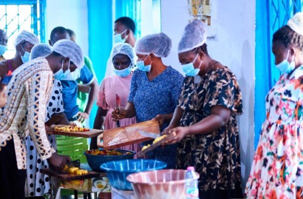 Screenshot 2026-05-21 083828 | Ghanaguardian.com People wearing masks and hairnets line up to receive food from a buffet-style table in a bright blue room.