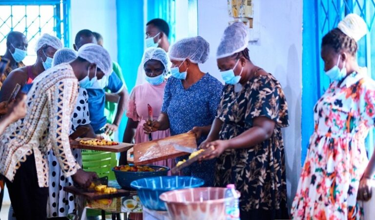 People wearing masks and hairnets line up to receive food from a buffet-style table in a bright blue room.
