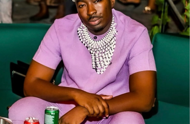 Man in a lavender outfit with a chunky beaded necklace sits on a green sofa at a party, with red and green cans on a table nearby.