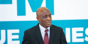 Man in a black suit and red striped tie speaks into a lapel microphone, set against a blue and white backdrop.
