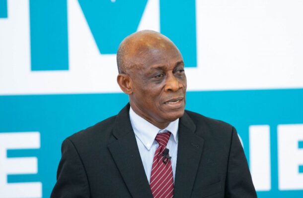 Man in a black suit and red striped tie speaks into a lapel microphone, set against a blue and white backdrop.