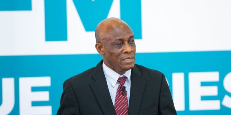 Man in a black suit and red striped tie speaks into a lapel microphone, set against a blue and white backdrop.