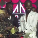 Man in a white outfit receives a star-shaped trophy from a woman with a gold headwrap on a crowded stage with a red patterned backdrop.