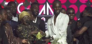 Man in a white outfit receives a star-shaped trophy from a woman with a gold headwrap on a crowded stage with a red patterned backdrop.