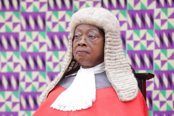 Female judge wearing a traditional white barrister's wig and red robes, seated with a serious expression, against a colorful geometric backdrop.