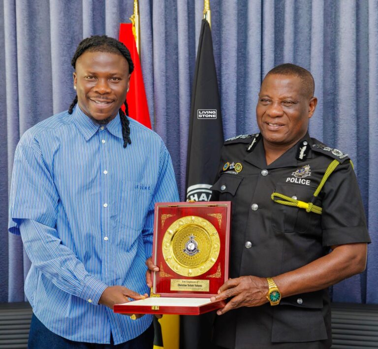 Man in a blue striped shirt and a police officer pose with a large gold-award plaque in a red frame, against blue curtains with flags behind them.