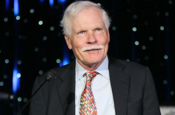 Older man with white hair and mustache in a dark suit, speaking at a podium microphone on a stage.