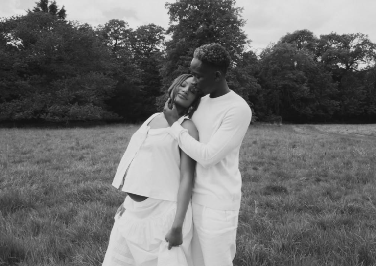 A couple stands in an open field, the man embracing the woman from side as they lean their heads together in a tender moment. Both dressed in white in a black-and-white photo with trees in the background.