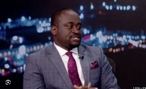 Man in a gray checkered suit with white shirt and maroon tie sits on a talk-show set, city lights backdrop and a lapel mic visible.