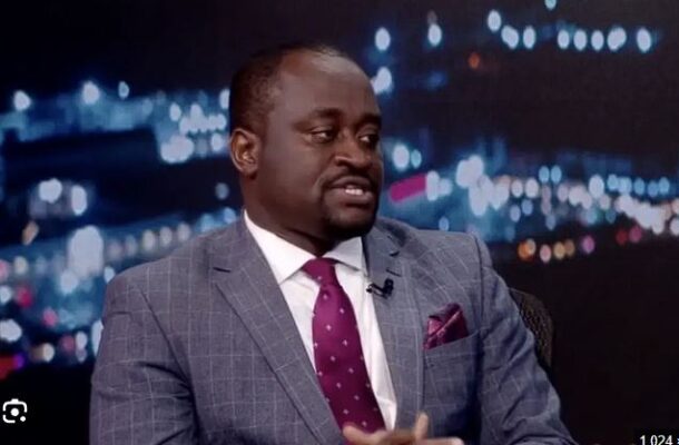Man in a gray checkered suit with white shirt and maroon tie sits on a talk-show set, city lights backdrop and a lapel mic visible.