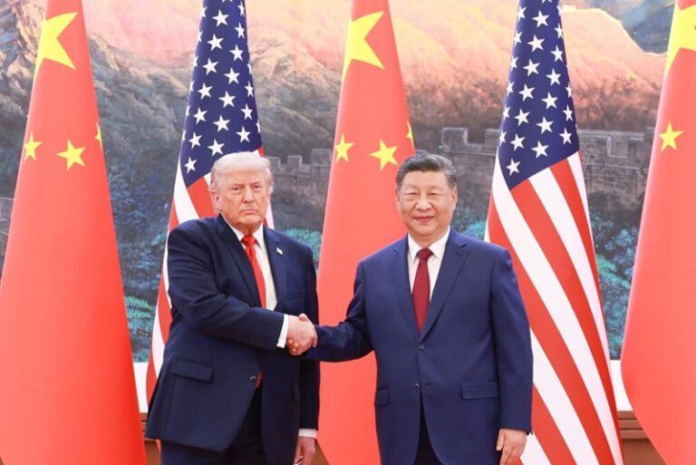 Two men in suits shake hands in front of US and China flags with a mountainous backdrop, signaling a diplomatic meeting.