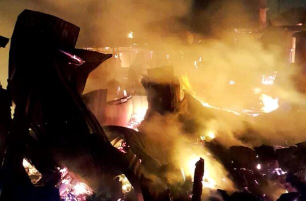 Nighttime scene of a large fire in a debris-filled area, orange flames and thick smoke, with an official-looking logo in the upper right corner.