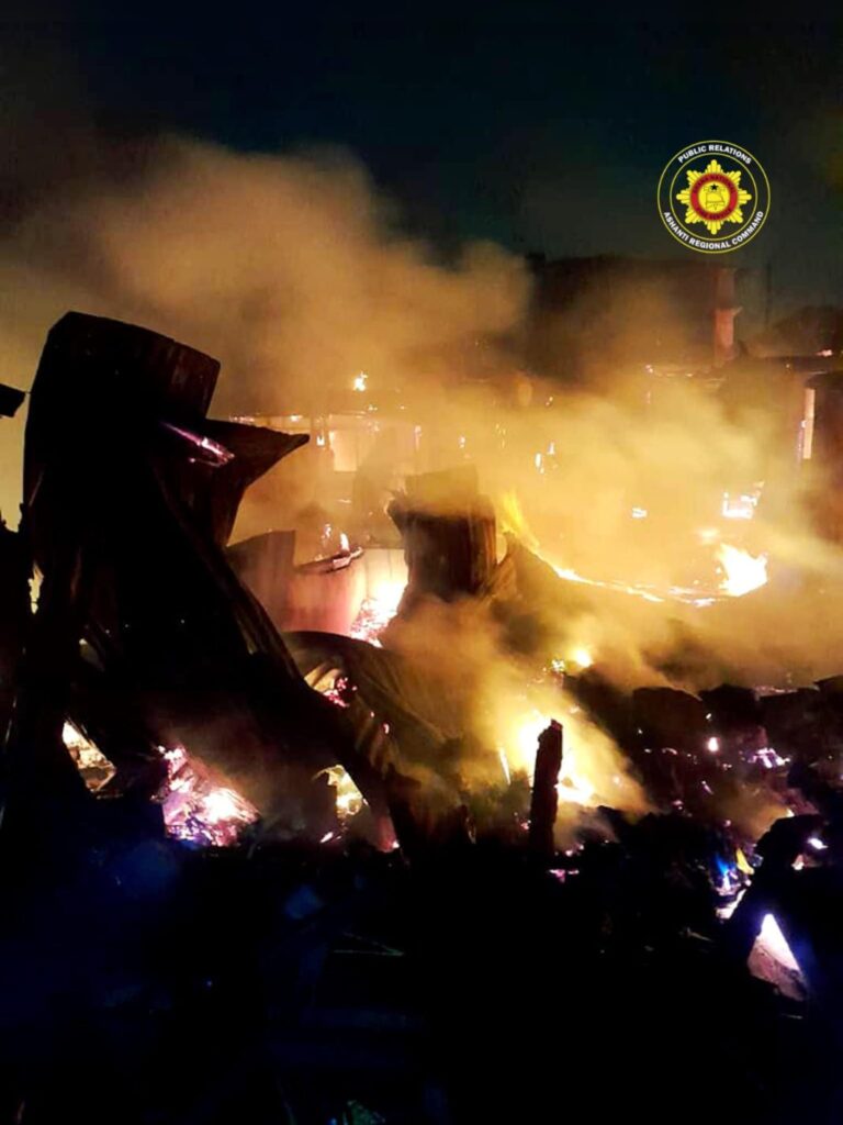 Nighttime scene of a large fire in a debris-filled area, orange flames and thick smoke, with an official-looking logo in the upper right corner.