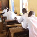 Students in white shirts sit at wooden desks in a sunlit classroom, focused on papers on their desks.