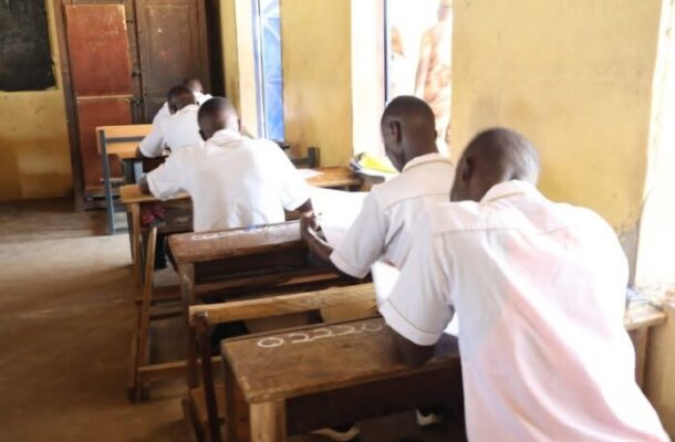 Students in white shirts sit at wooden desks in a sunlit classroom, focused on papers on their desks.