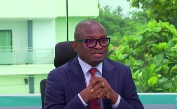 WhatsApp-Image-2025-11-27-at-42354-AM-750x375 | Ghanaguardian.com Man in a navy suit with red tie and glasses sits at a desk with hands clasped, in a studio with greenery outside.