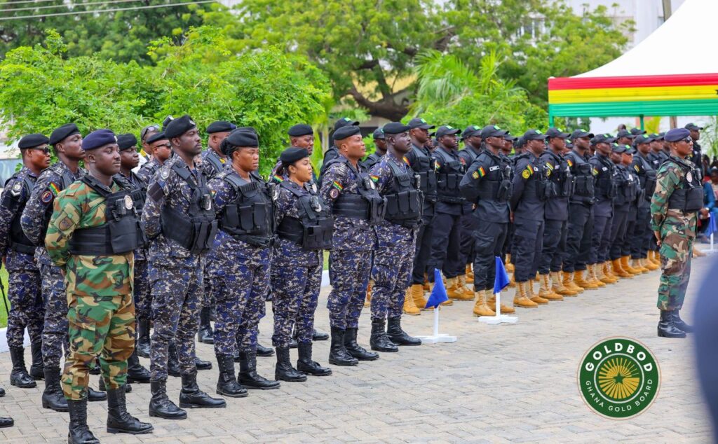 Line of uniformed security personnel standing at attention in a ceremonial parade, with camouflage and dark uniforms and a rainbow-striped tent in the background.