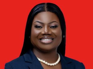 Professional headshot of a smiling Black woman in a navy blazer and pearl necklace against a solid red background.