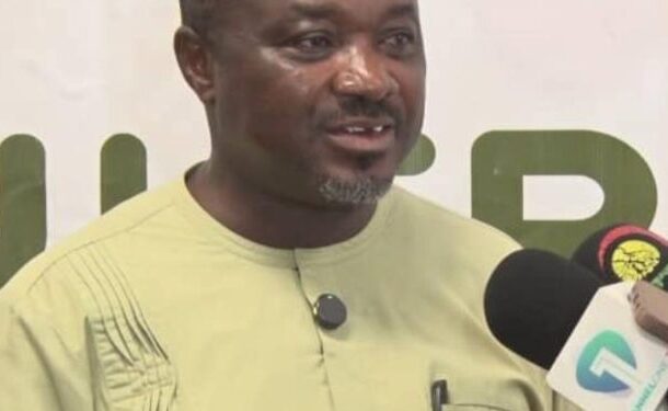 Man in a light green traditional outfit speaks into a microphone during an interview.