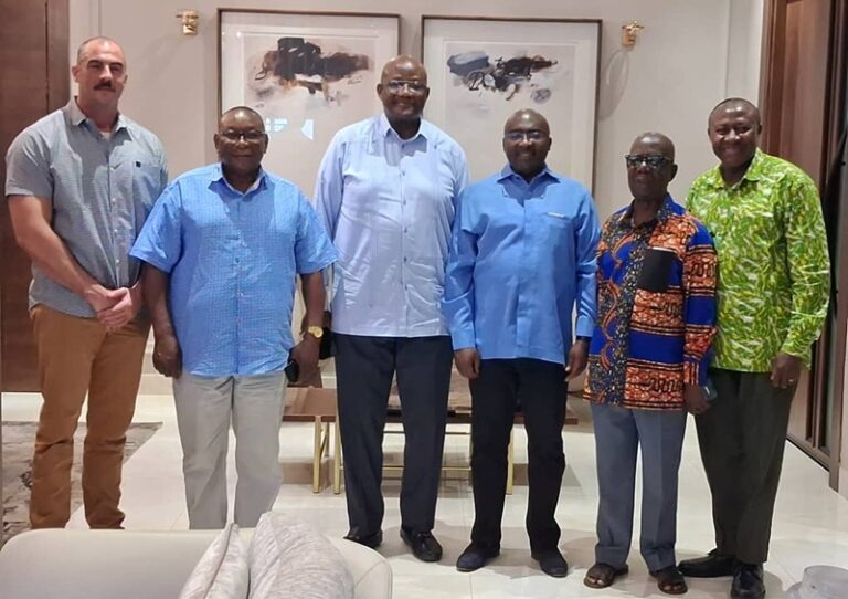 Six men stand side by side indoors, posing for a group photo in a hallway or reception area.